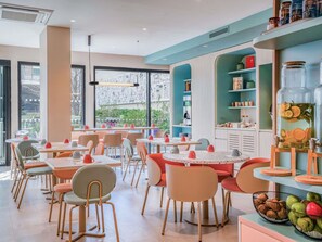 A modern dining area with round tables and chairs, a large window with a view, and a kitchen area with shelves and jars. - ibis Styles Paris Porte d’Orléans Hotel (Montrouge)