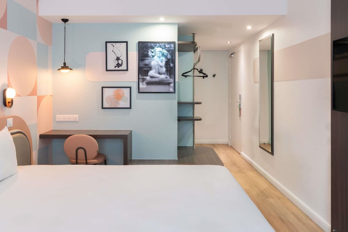 A modern hotel room with a bed, a desk, a chair, a wardrobe, and artwork on the walls.