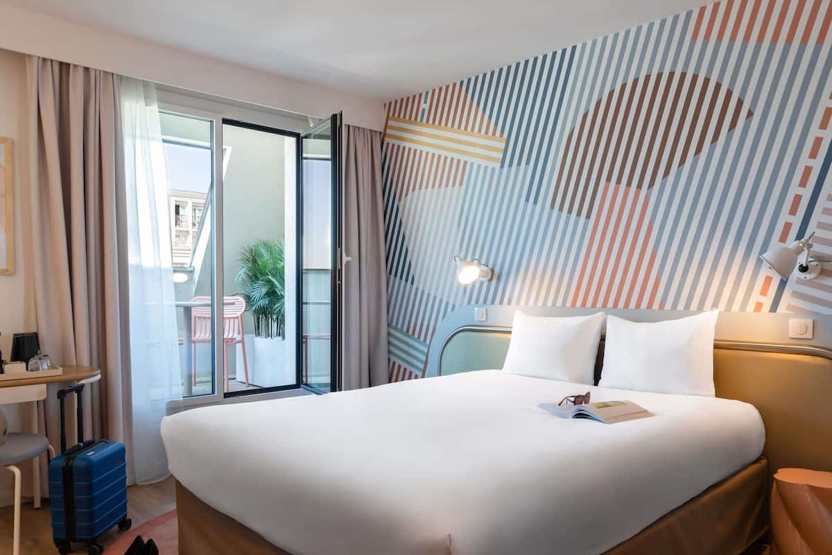 A hotel room with a bed, a desk, a chair, a suitcase, and a view of the outside.