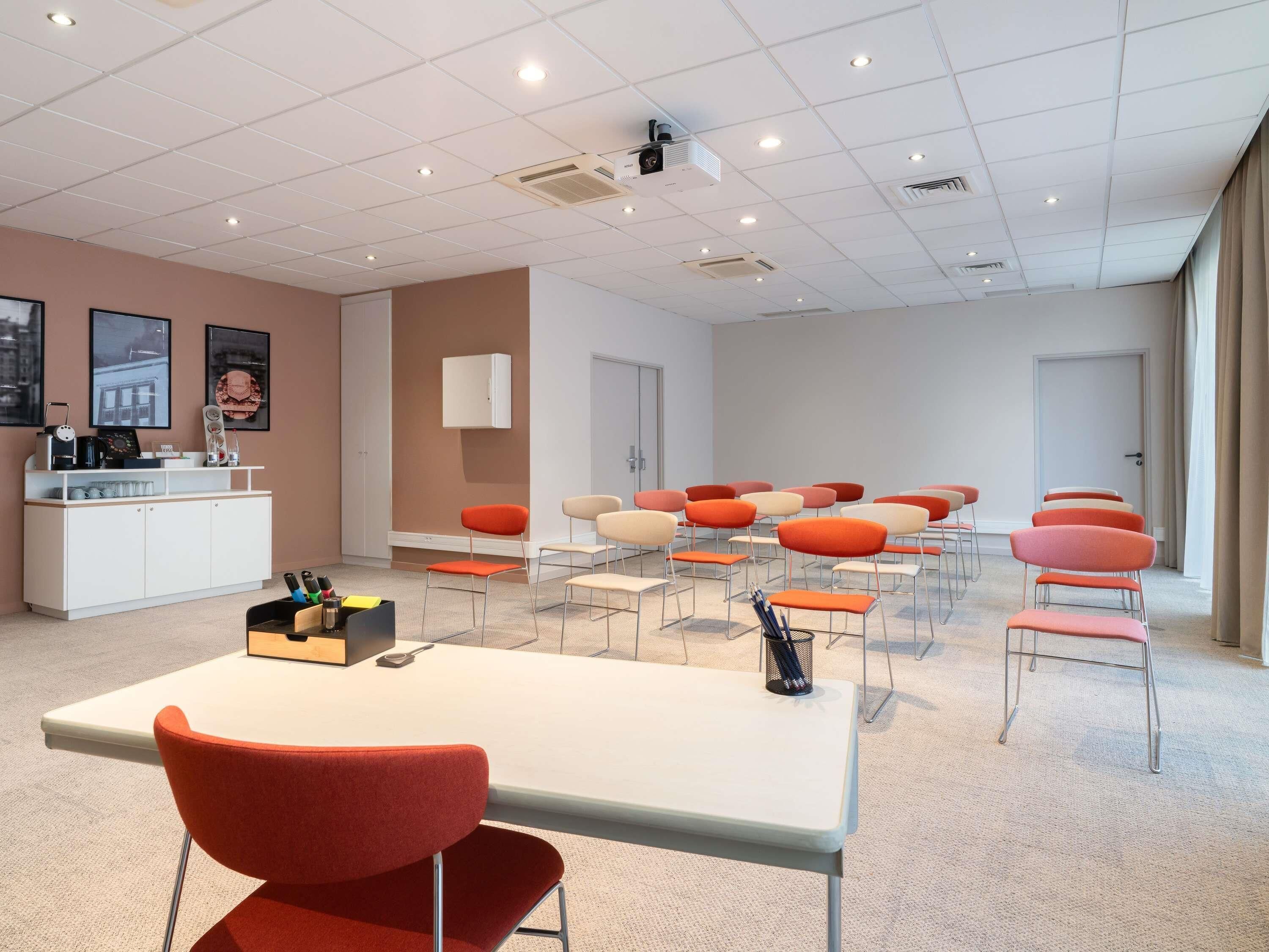 Une salle de conférence avec une table blanche, des chaises rouges et beige, un meuble blanc et un projecteur fixé au plafond.