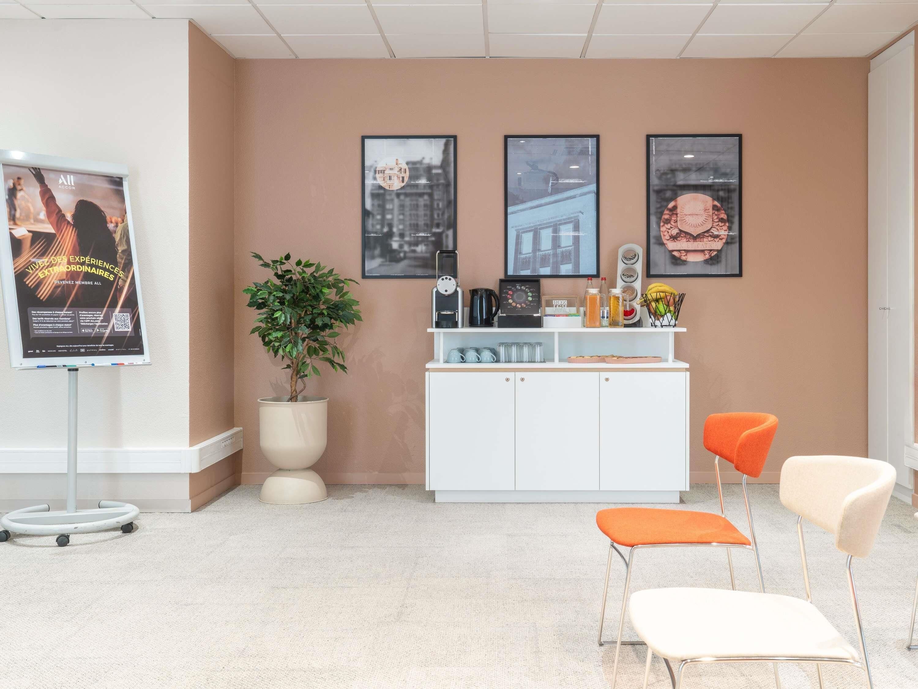 A reception area with a white counter, a potted plant, framed pictures on the wall, and two orange chairs.