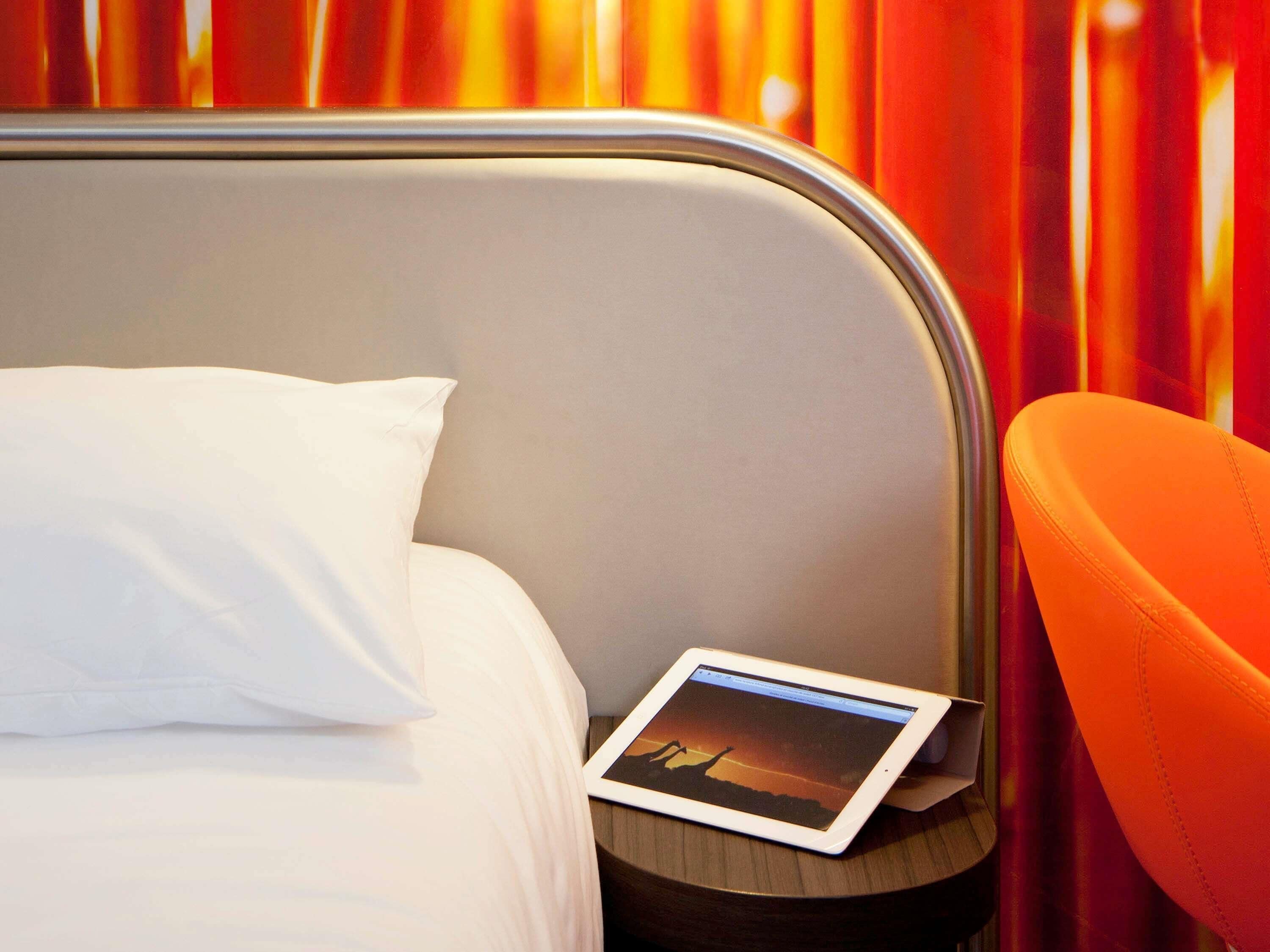 Un lit avec un oreiller blanc, une tablette posée sur la table de chevet et une chaise orange.