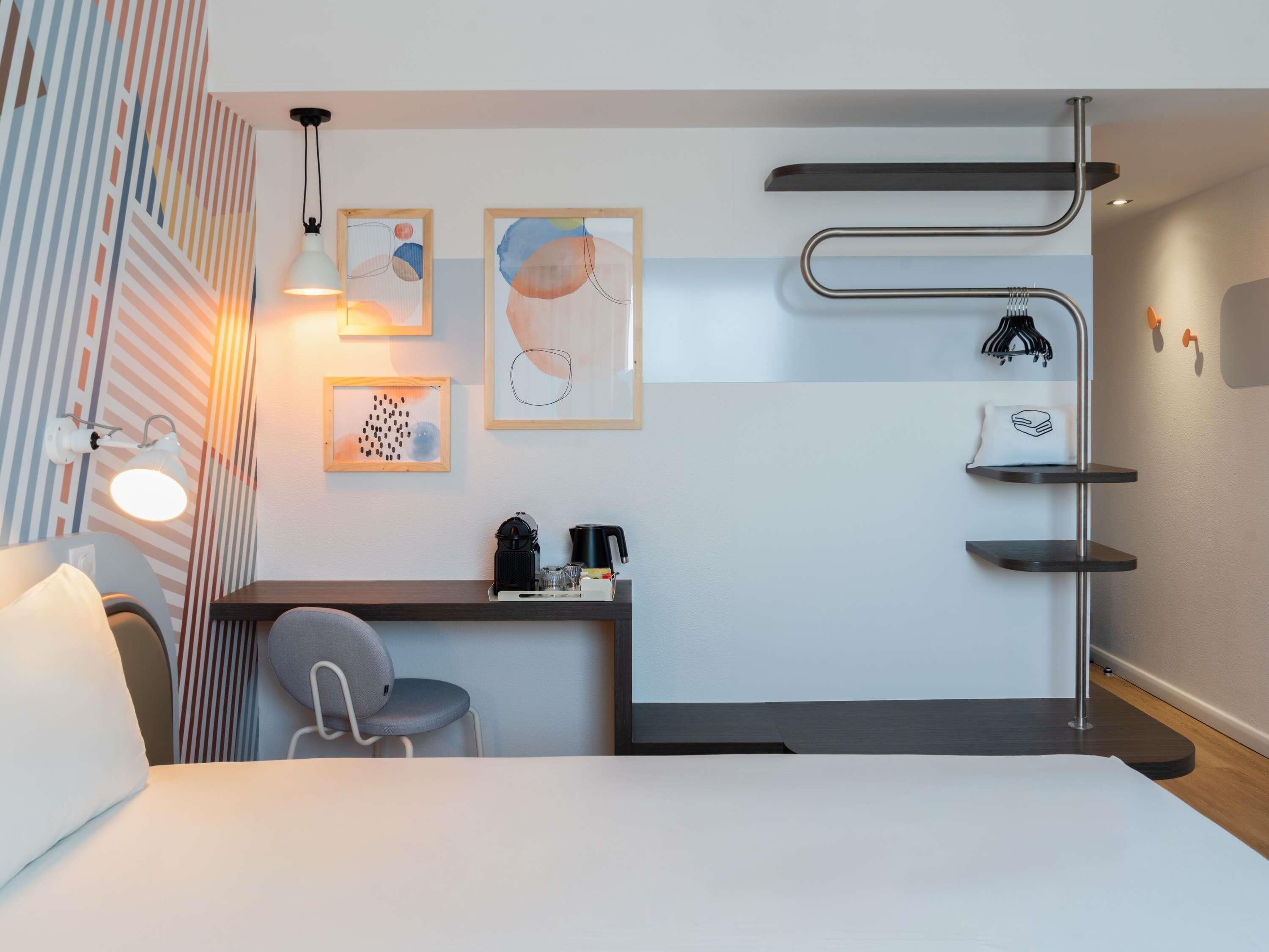 A modern hotel room with a bed, a desk, a chair, a coffee maker, and wall-mounted shelves.