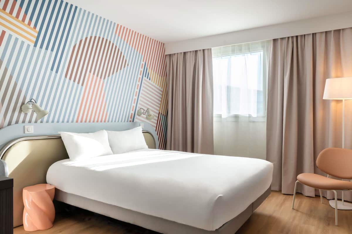 A modern hotel room with a large bed, a bedside table, a chair, and a wall with a striped pattern.