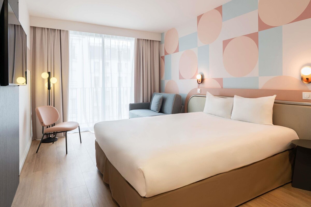 A hotel room with a large bed, a seating area with a chair, a sofa, and a wall-mounted light fixture.