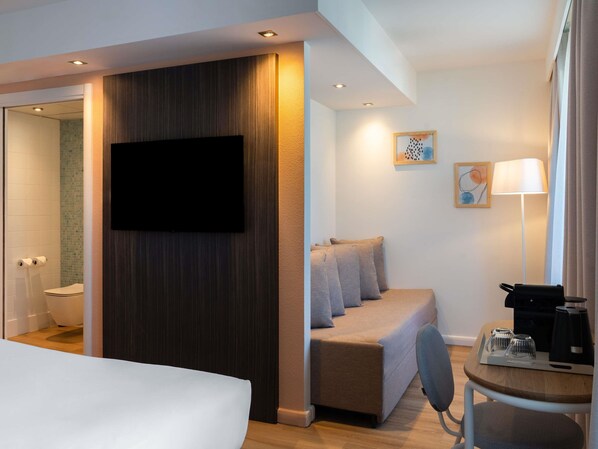 A modern hotel room with a bed, a sofa, a desk with a coffee maker, and a TV mounted on the wall. - ibis Styles Paris Porte d’Orléans Hotel (Montrouge)