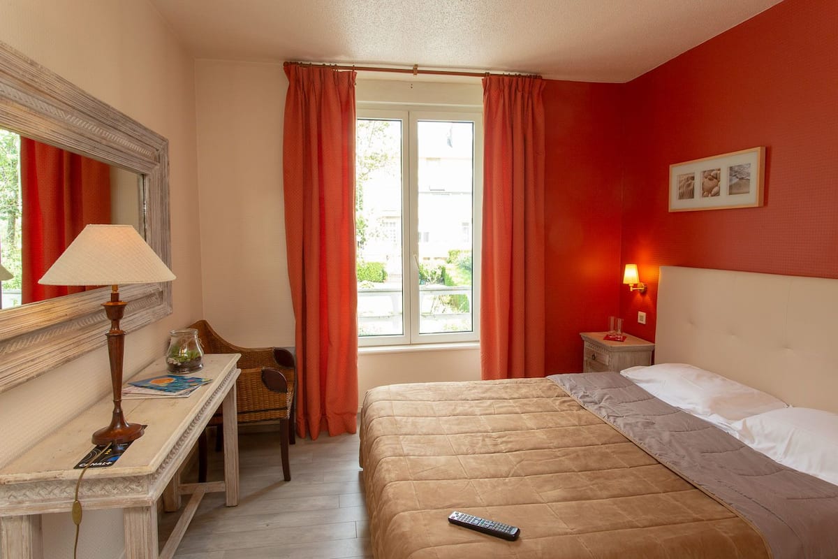 A hotel room with a bed, a desk with a lamp, a chair, a nightstand with a lamp, and a window with curtains.