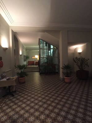 A hallway with patterned flooring, potted plants, and a staircase leading to a room with a bed and a lamp. - Hôtel Providence (Vittel)