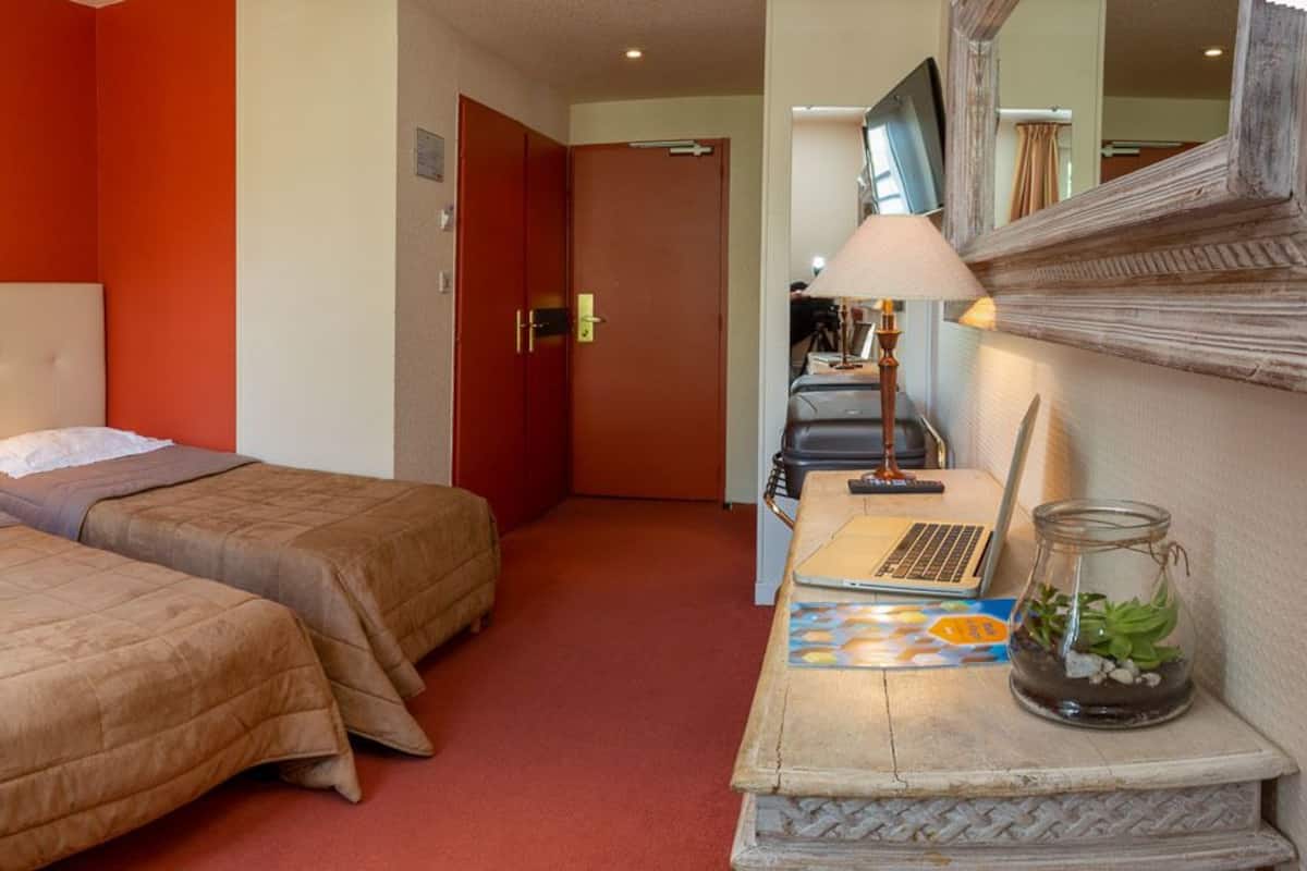 A hotel room with two beds, a desk with a laptop, a lamp, and a potted plant.