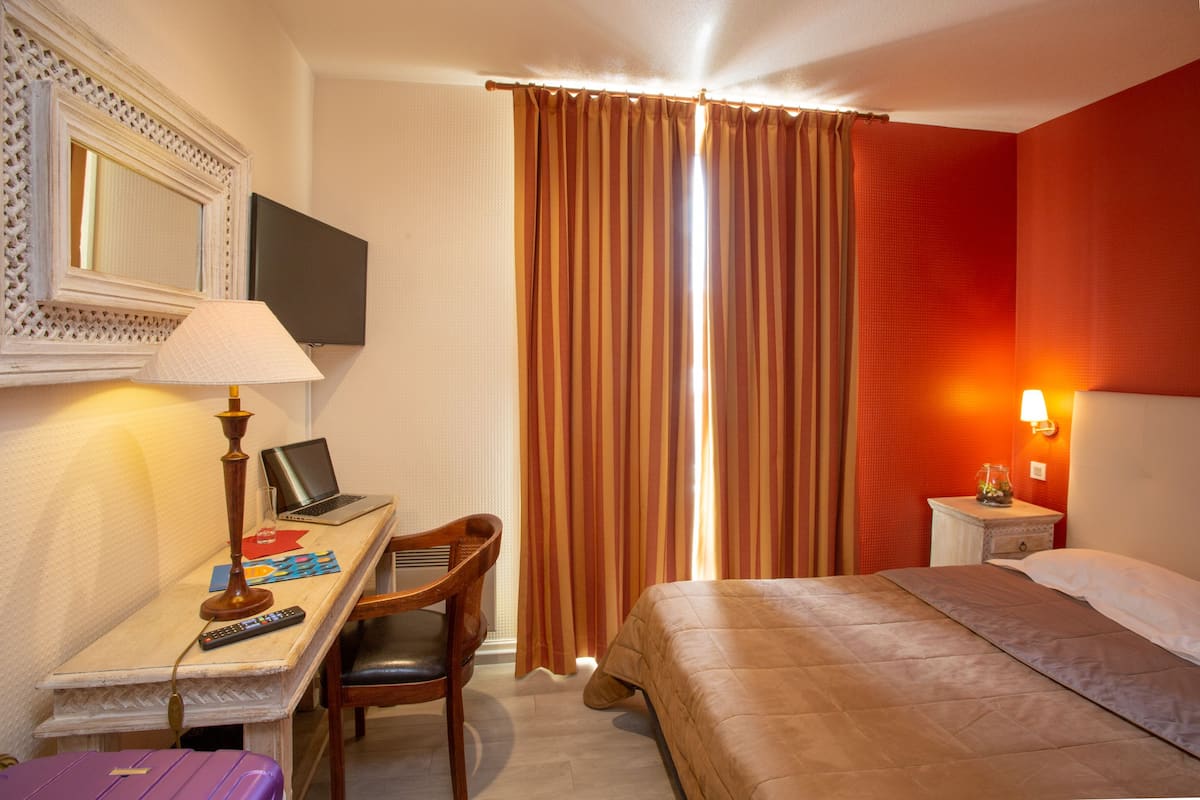 A hotel room with a bed, a desk with a laptop, a chair, a lamp, a TV, and a window with curtains.