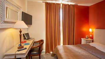 A hotel room with a bed, a desk with a laptop, a chair, a lamp, a TV, and a window with curtains.