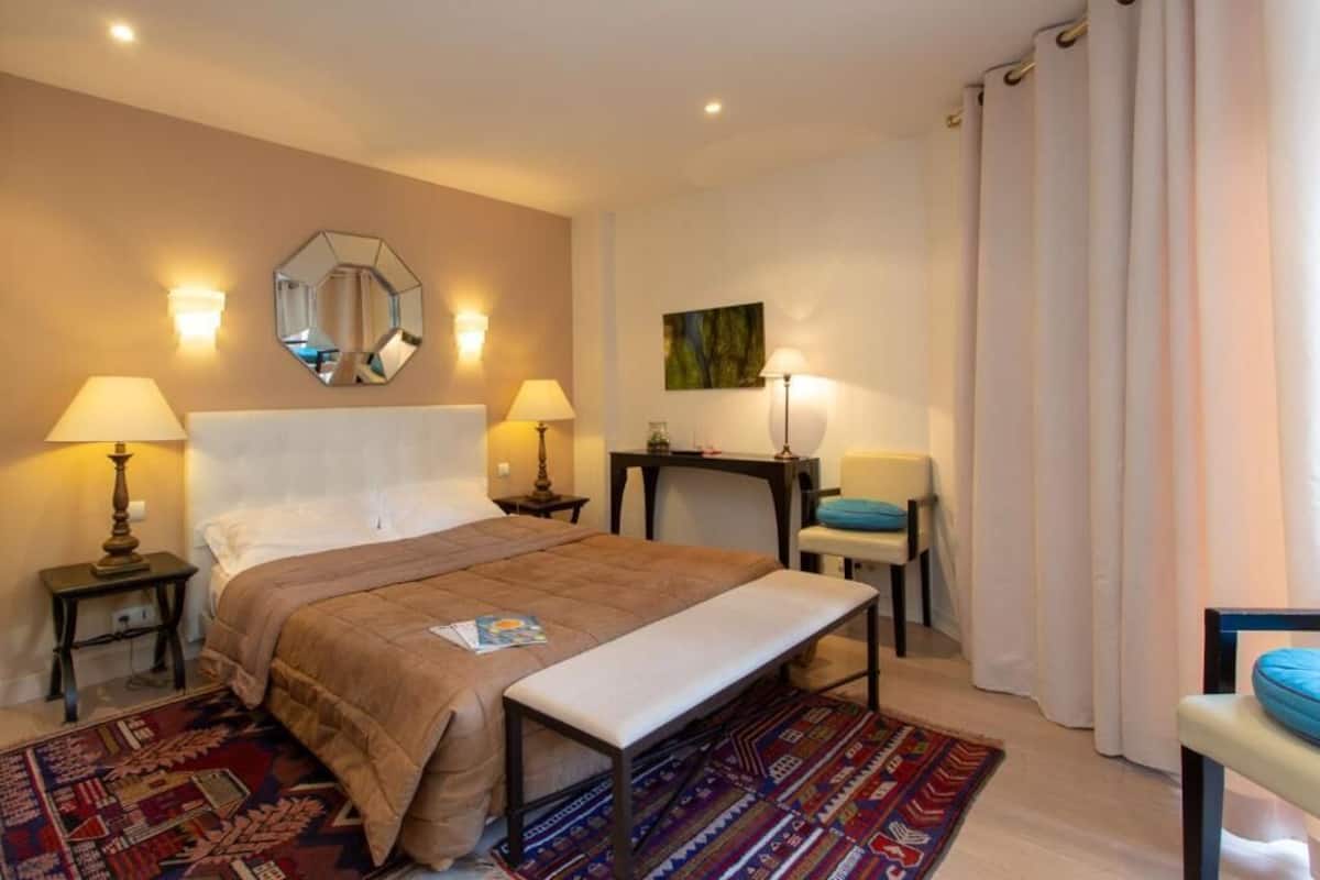A hotel room with a bed, bedside lamps, a desk, a chair, a bench, a rug, a mirror, and curtains.