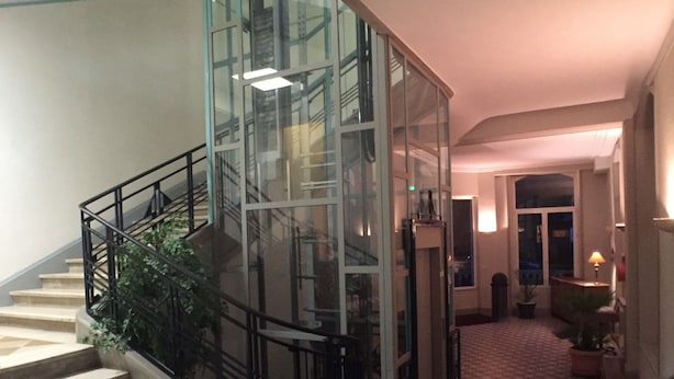 A staircase with glass railings leading to an upper level, a checkered floor, and a hallway with a lamp and potted plant. - Hôtel Providence (Vittel)