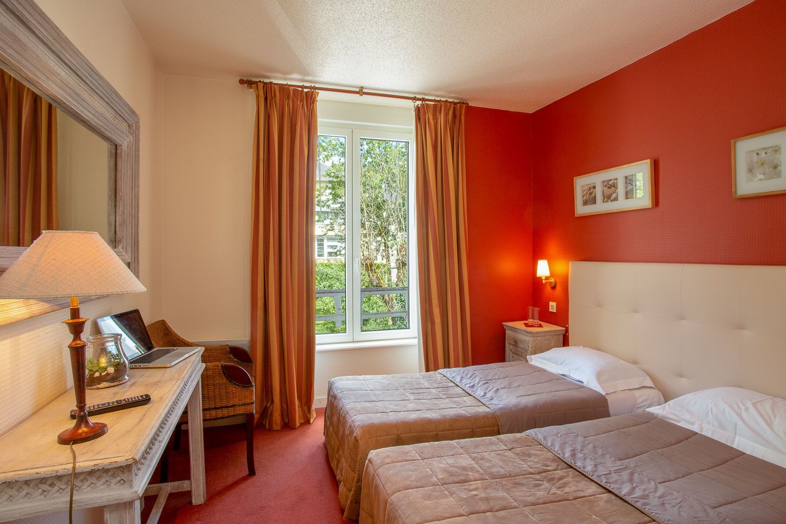 Habitación de hotel con dos camas, un escritorio con lámpara, una silla, una ventana con cortinas y una mesita de noche con lámpara.
