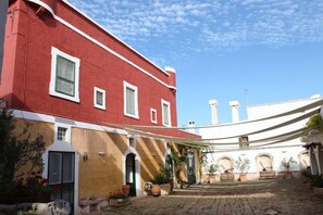 A courtyard with a red building, a yellow wall, and a white building with arches. There are potted plants and a bench. - Masseria Refrigerio (Ostuni)