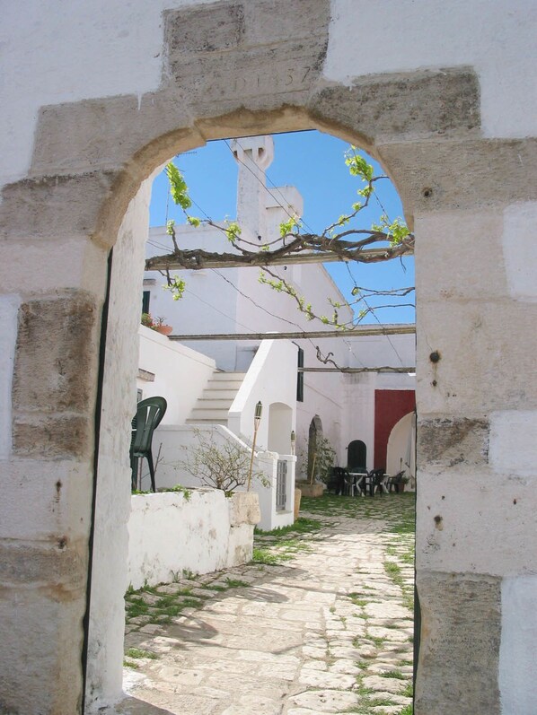 An arched doorway leading to a courtyard with a stone pathway, white walls, and a red building in the background. - Masseria Refrigerio (Ostuni)