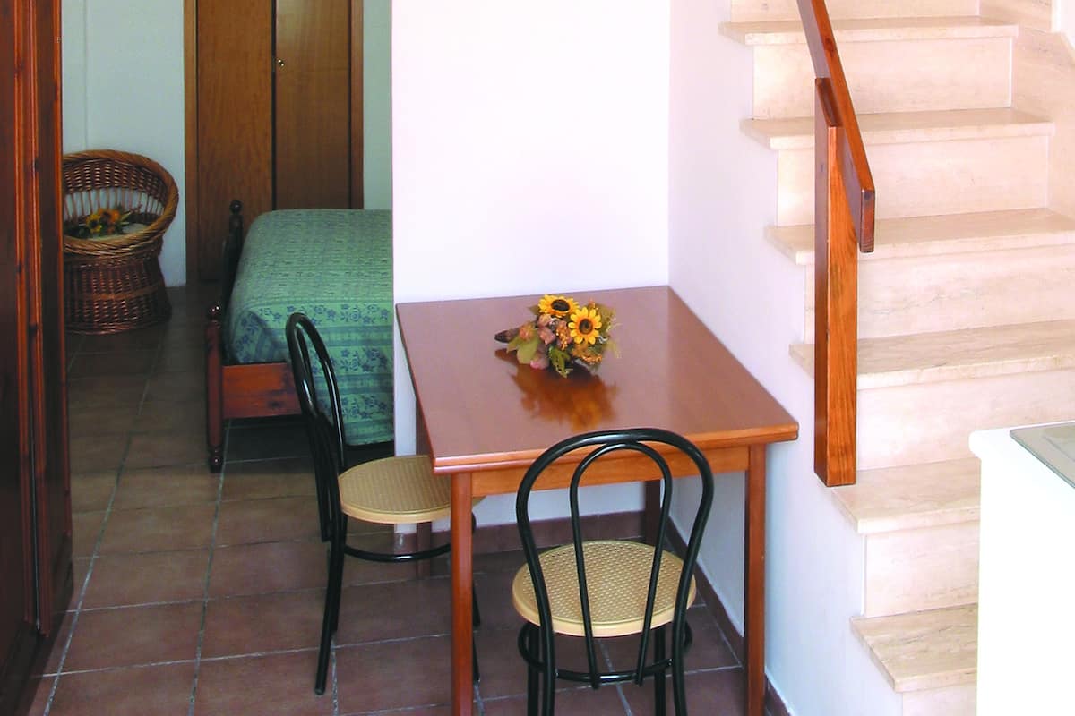 A small room with a wooden table, two chairs, a bed, a wicker chair, and a staircase.