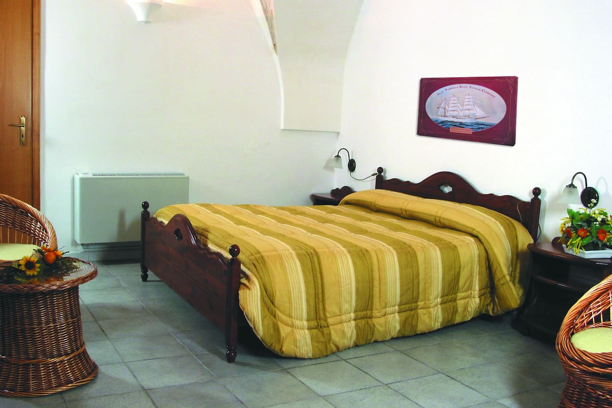 A bedroom with a bed, a wicker chair, a nightstand, a wall-mounted lamp, and a framed picture.