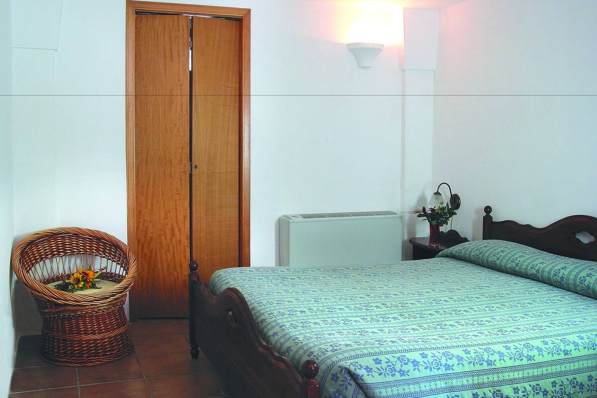 A bedroom with a wooden wardrobe, a wicker chair, a bedside table with a lamp, and a bed with patterned bedding.