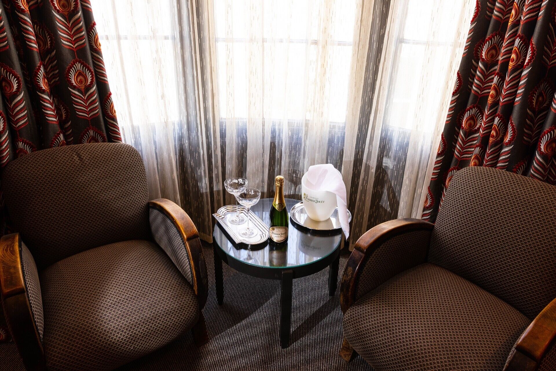 A room with two upholstered chairs, a small round table with a bottle of champagne, a glass, and a white bowl.
