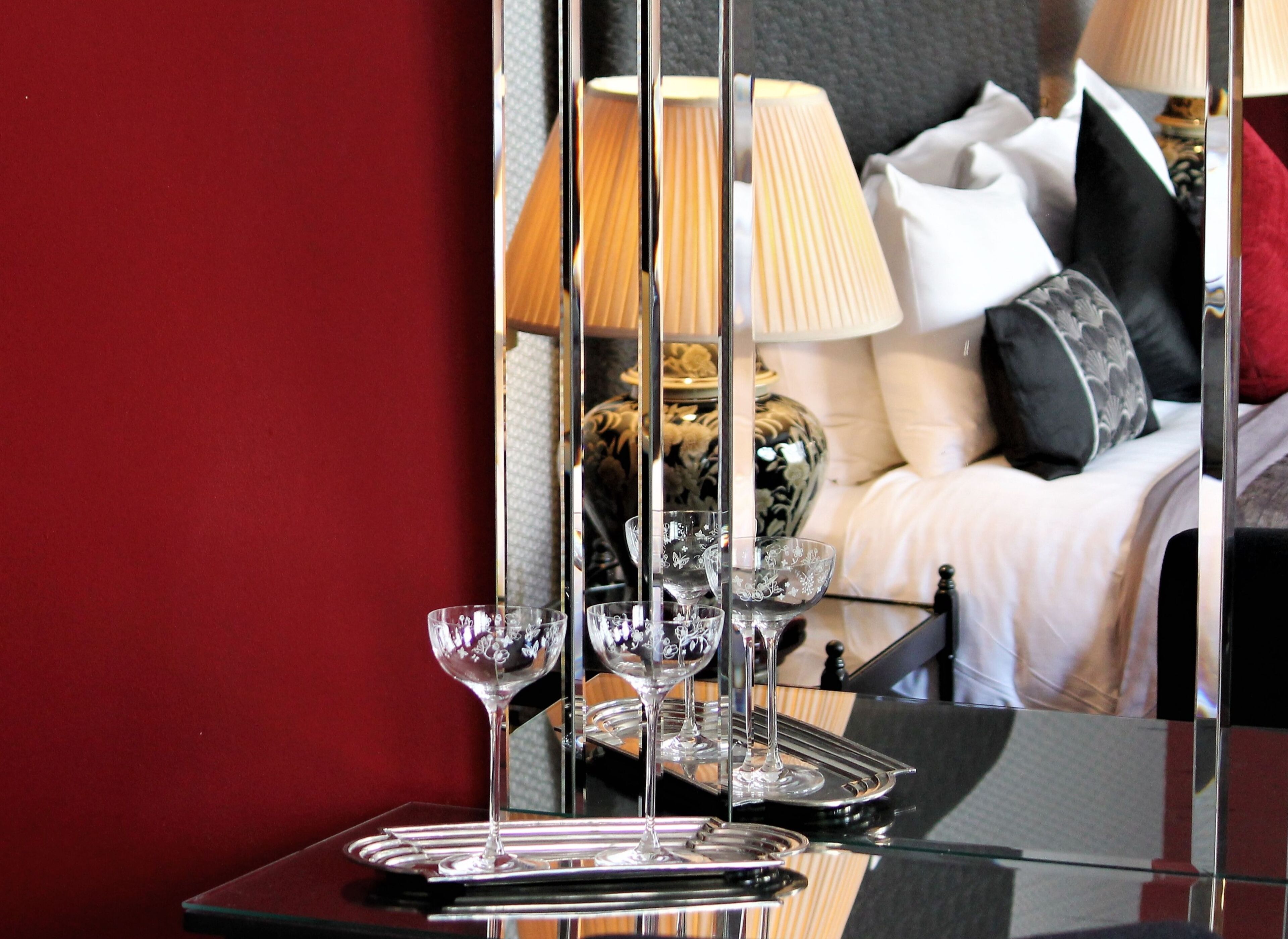 A glass table with wine glasses and a tray, reflecting a room with a bed, pillows, and a lamp.