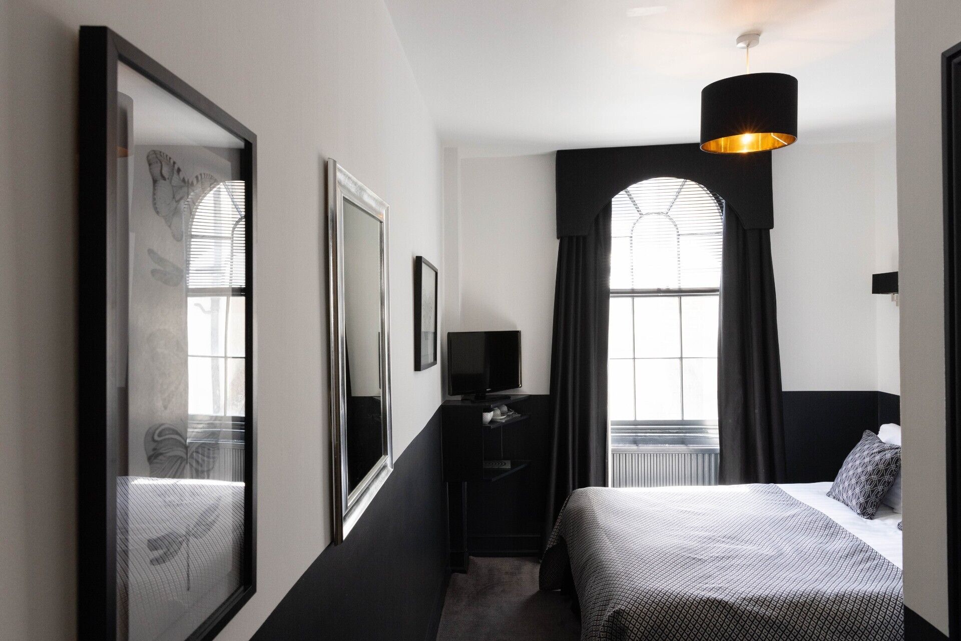 A modern hotel room with a large bed, a television, and a mirror. There is a window with curtains and a black lamp hanging from the ceiling.