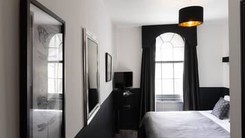 A modern hotel room with a large bed, a television, and a mirror. There is a window with curtains and a black lamp hanging from the ceiling.