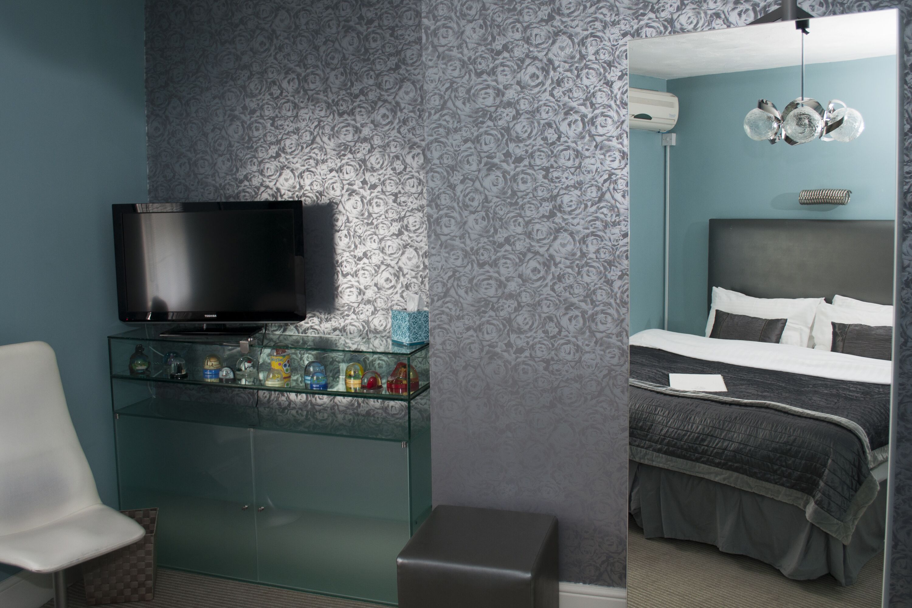A modern hotel room with a flat-screen TV on a glass shelf, a bed with a dark headboard, and a patterned wall.