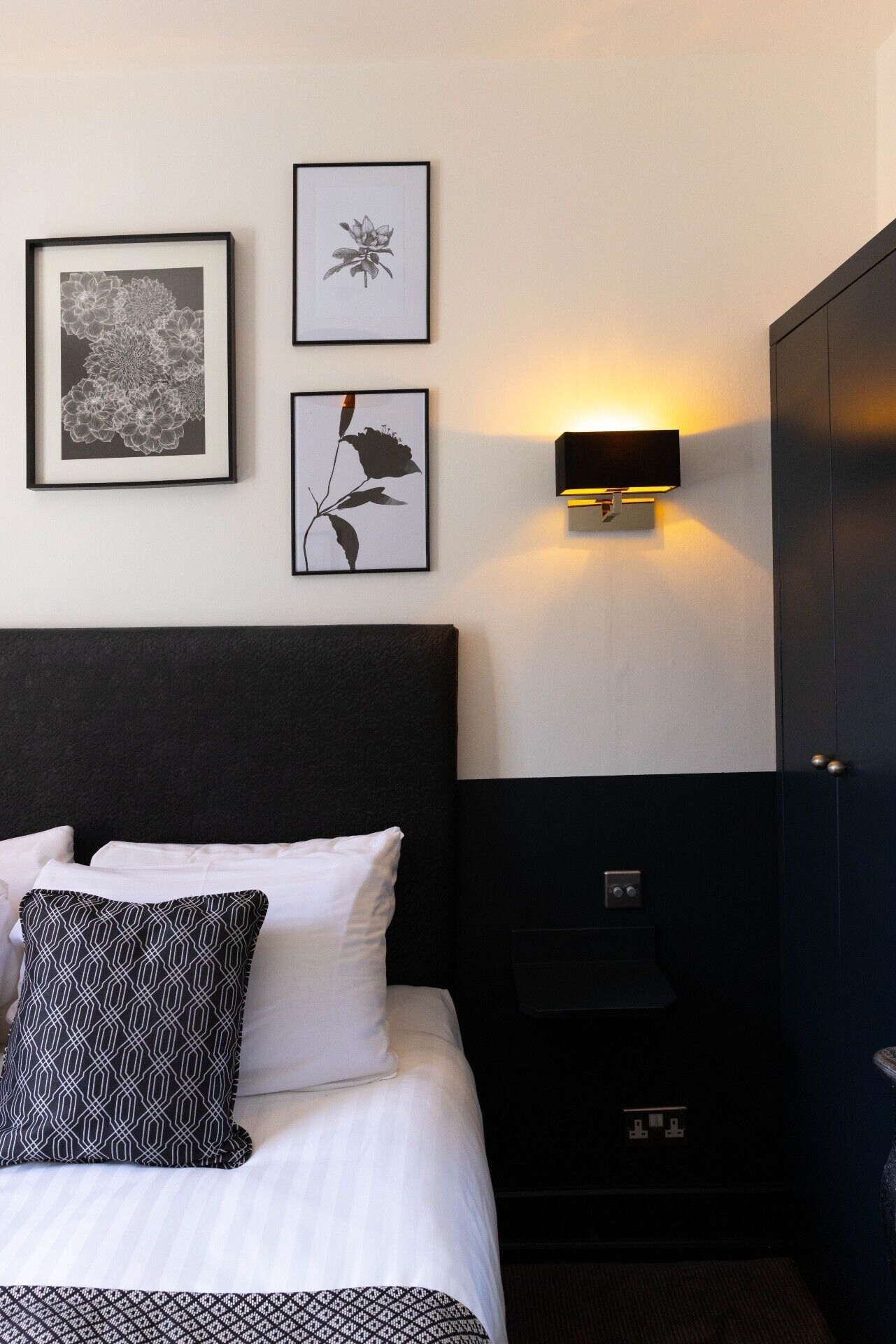 Une chambre d’hôtel avec une tête de lit noire, du linge de lit blanc et trois œuvres d’art florales encadrées accrochées au mur.