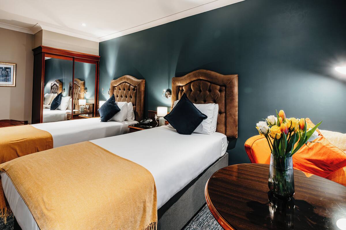 Une chambre d’hôtel avec deux lits, une tête de lit en bois, une table de chevet avec une lampe, un vase avec des fleurs et une table ronde en bois.
