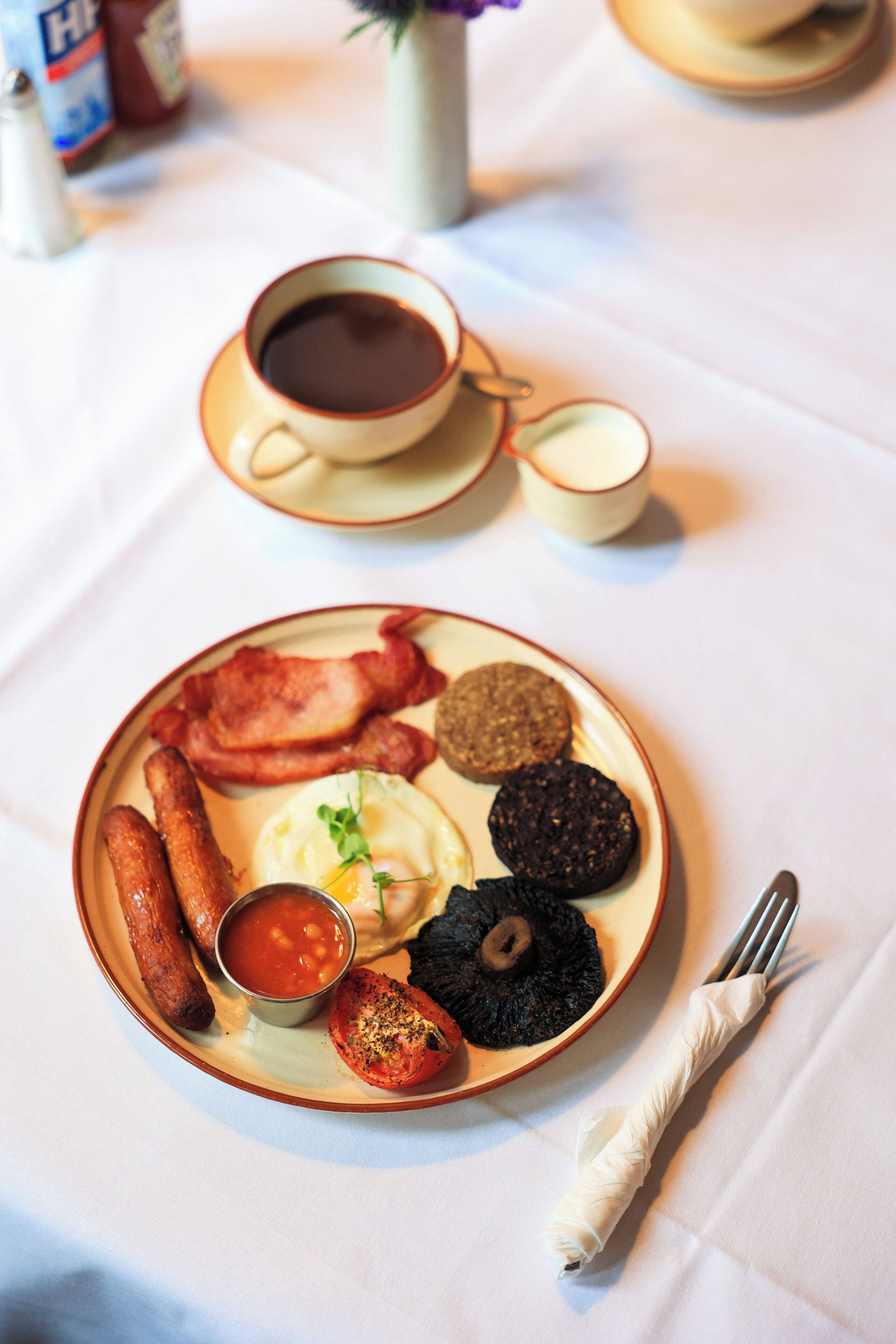 Un plato de desayuno con salchichas, huevos y morcilla, acompañado de una taza de café y un recipiente pequeño con salsa.