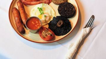 Un plato de desayuno con salchichas, huevos y morcilla, acompañado de una taza de café y un recipiente pequeño con salsa.