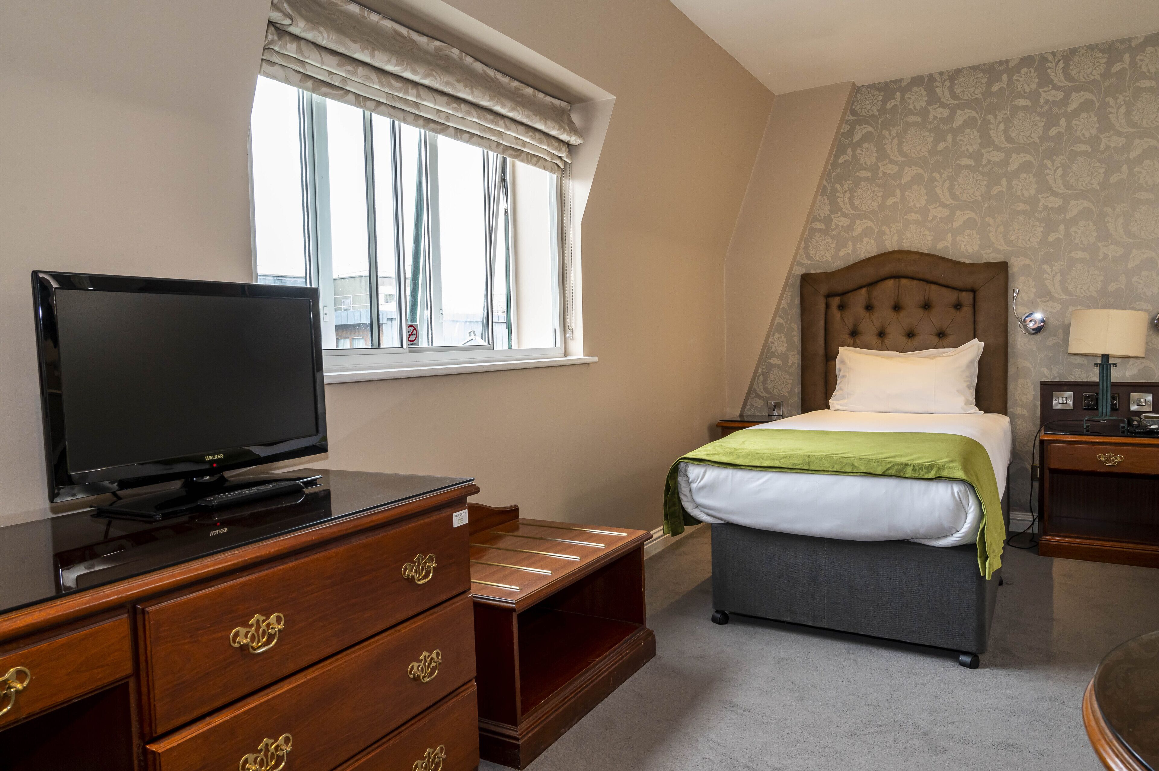 A hotel room with a bed, a television on a wooden dresser, a window with curtains, and a nightstand with a lamp.