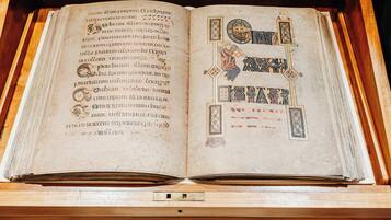 An open book with ornate designs and text, displayed in a wooden case with a glass front.