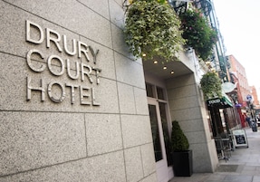 The exterior of the Drury Court Hotel with a stone facade, hanging plants, and a sign indicating a restaurant nearby.