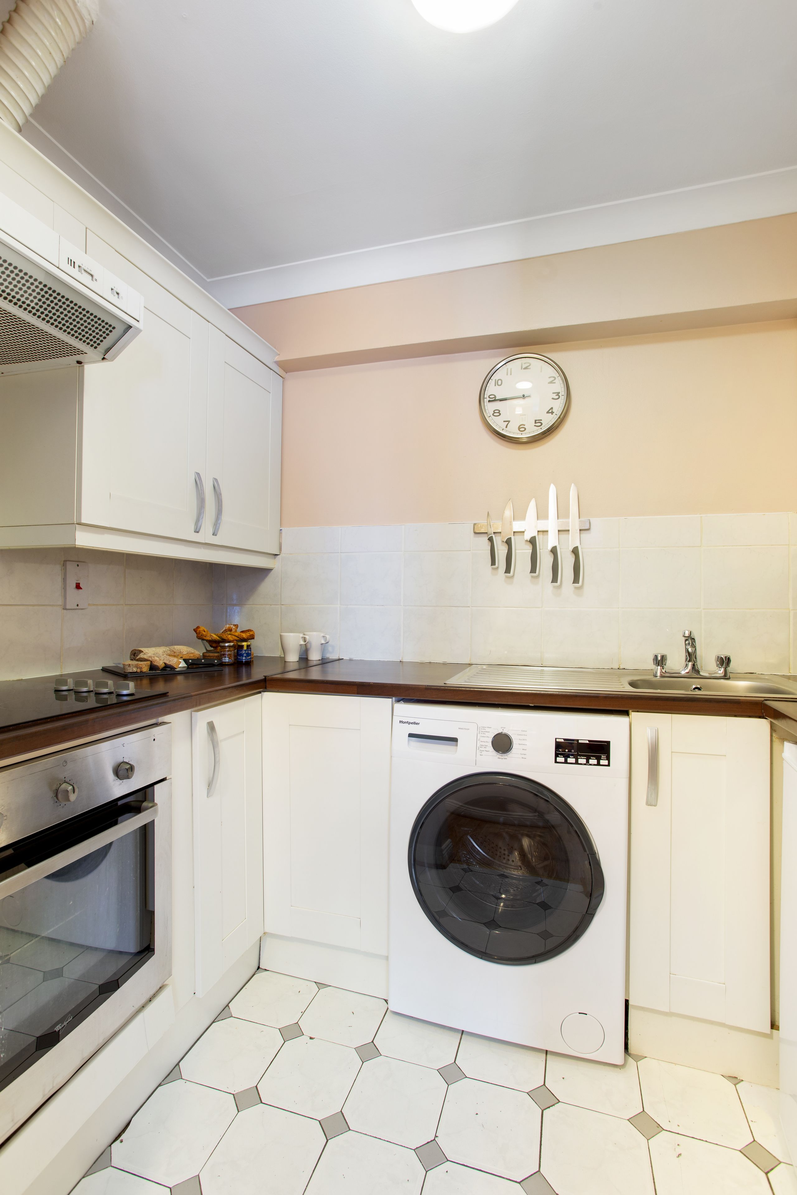 A compact kitchen with white cabinets, a built-in oven, a washing machine, and a clock on the wall.
