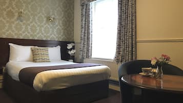 Habitación de hotel con una cama grande, una mesita de noche con un jarrón de flores, una ventana con cortinas estampadas y una pequeña mesa redonda con una tetera y una taza.