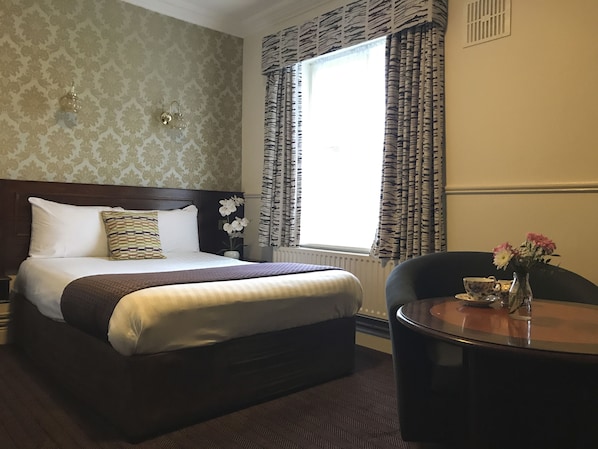 Habitación de hotel con una cama grande, una mesita de noche con un jarrón de flores, una ventana con cortinas estampadas y una pequeña mesa redonda con una tetera y una taza.