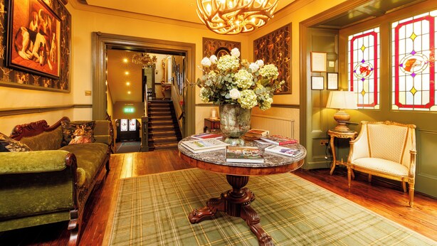 A well-lit living room with a chandelier, a green sofa, a round table with a vase of flowers, and a chair. - The Club House Hotel (Kilkenny)