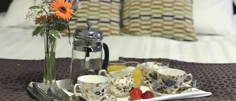 A breakfast tray with a coffee pot, teacups, a cup of orange juice, and strawberries on a bed with patterned bedding and pillows.