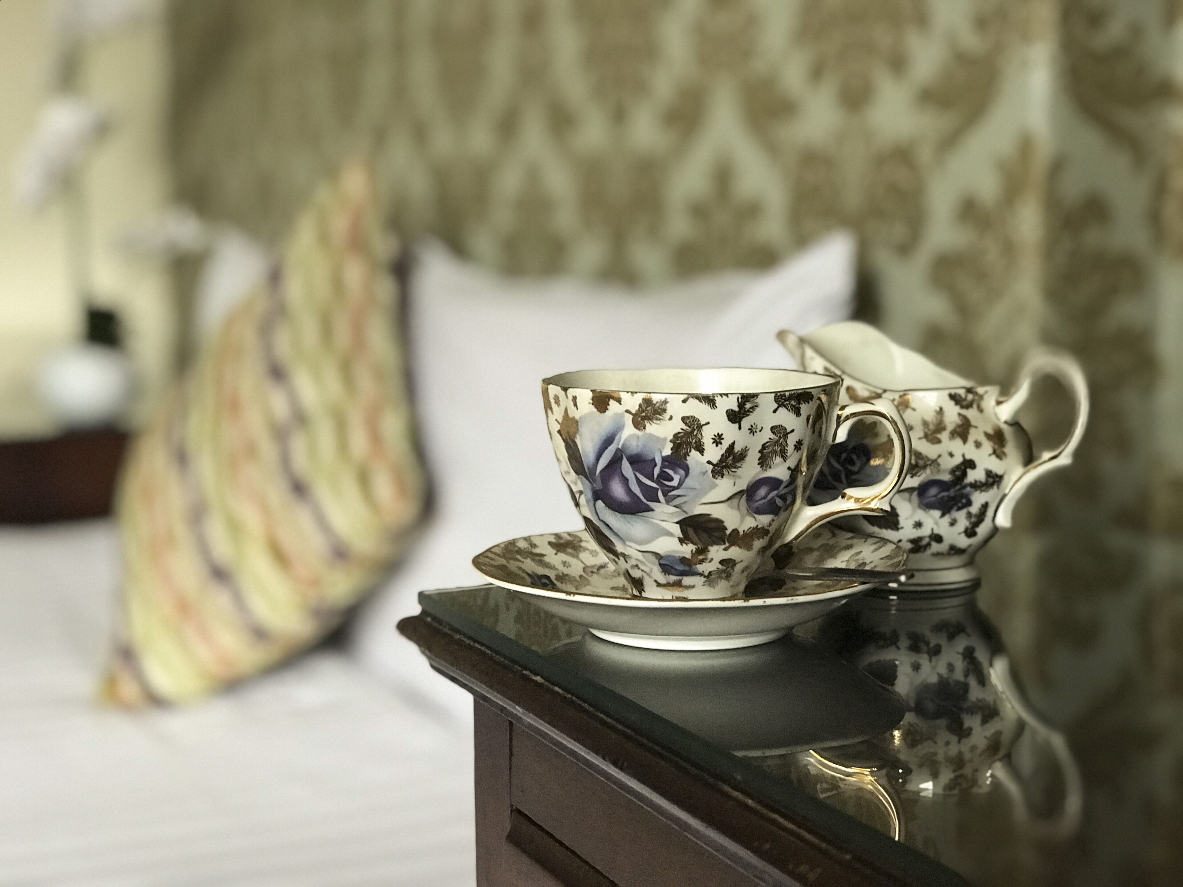 A tea set on a reflective surface, likely a table, with a patterned pillow and a bed in the background.