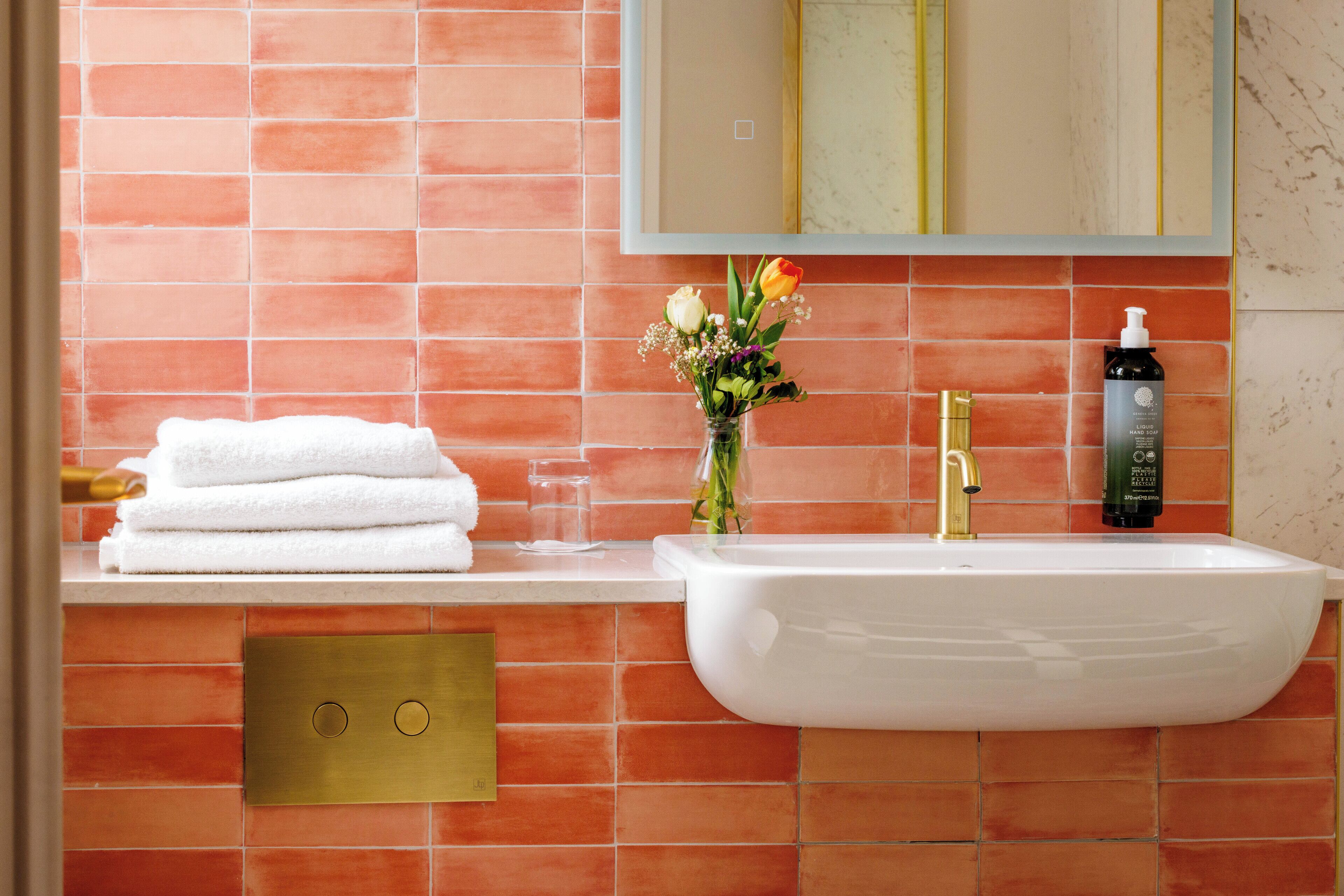 Un baño con un lavamanos blanco, un grifo dorado y un espejo encima. Hay azulejos naranja en la pared y un jarrón con flores en la encimera.