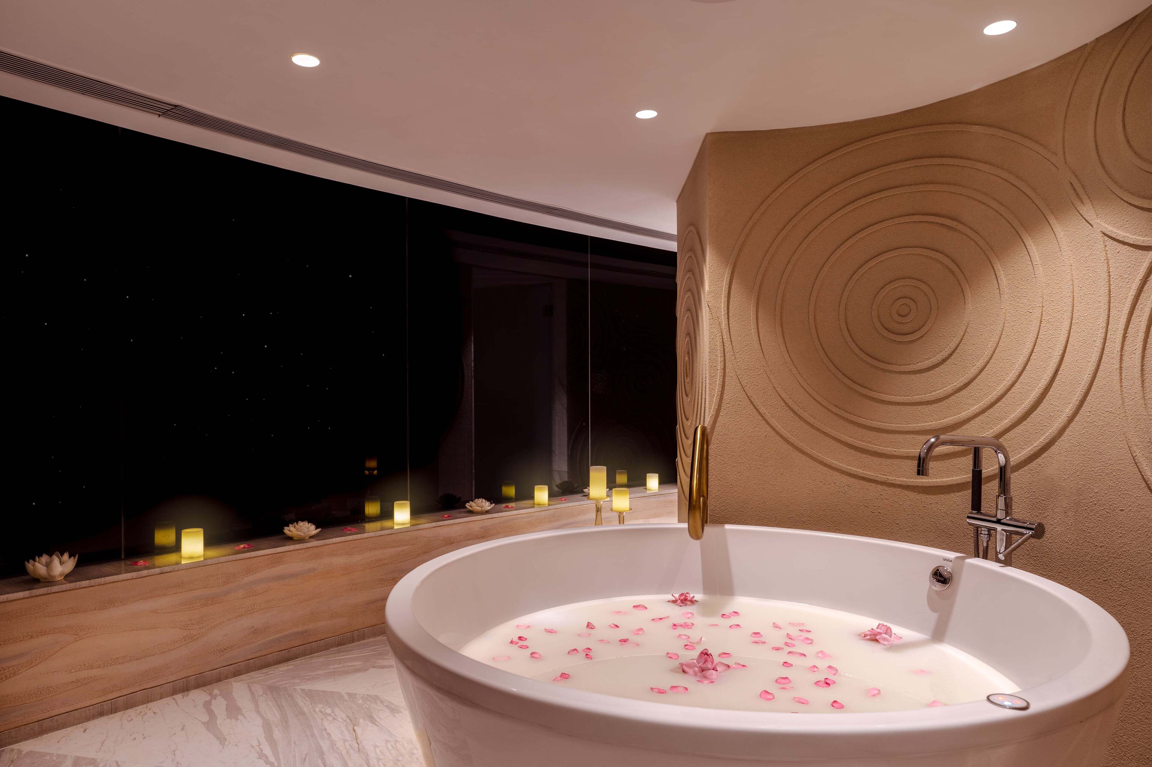 A modern bathroom with a large bathtub filled with water and rose petals, a wooden bench, and a wall with circular patterns.