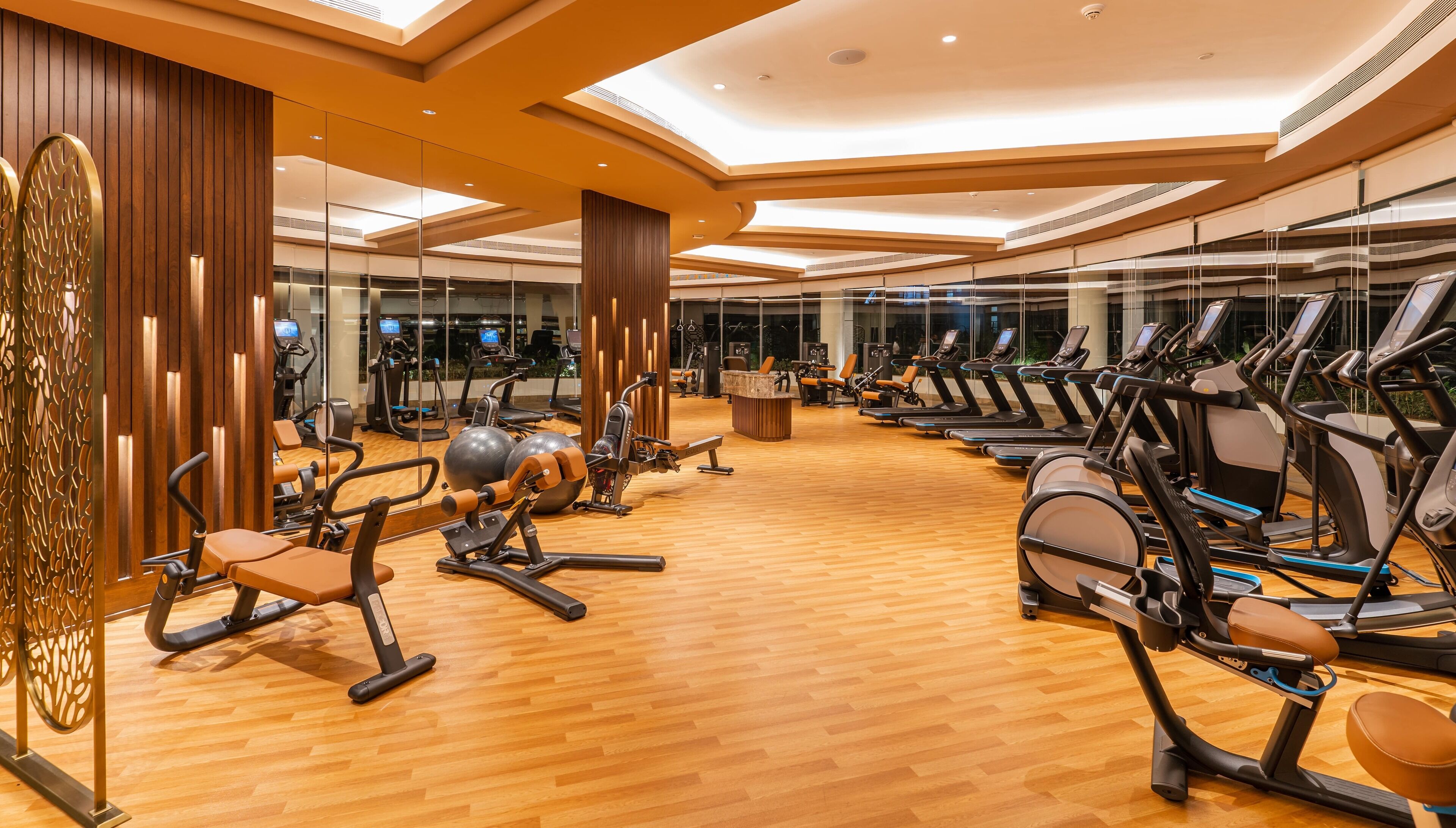 A modern gym with various exercise equipment, wooden flooring, and large windows.