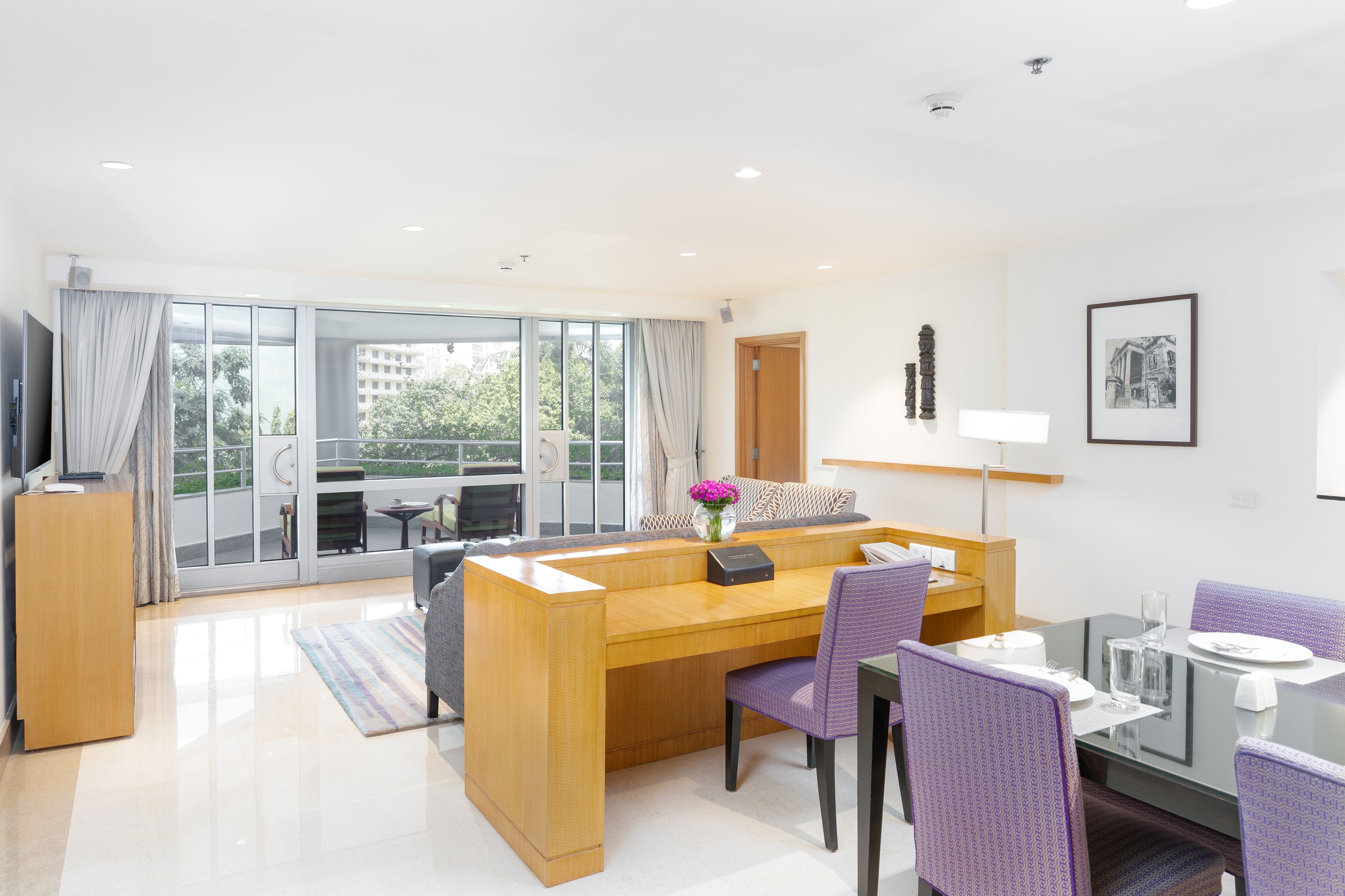 A modern hotel room with a wooden desk, purple chairs, a glass dining table, and a large window with a view of buildings and trees.