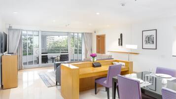 A modern hotel room with a wooden desk, purple chairs, a glass dining table, and a large window with a view of buildings and trees.