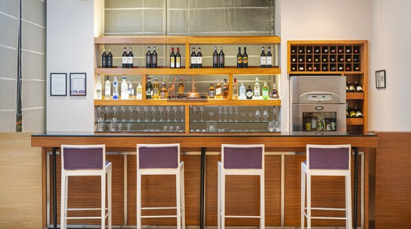 Una zona de bar con varias estanterías llenas de botellas y vasos, un refrigerador y taburetes de bar.