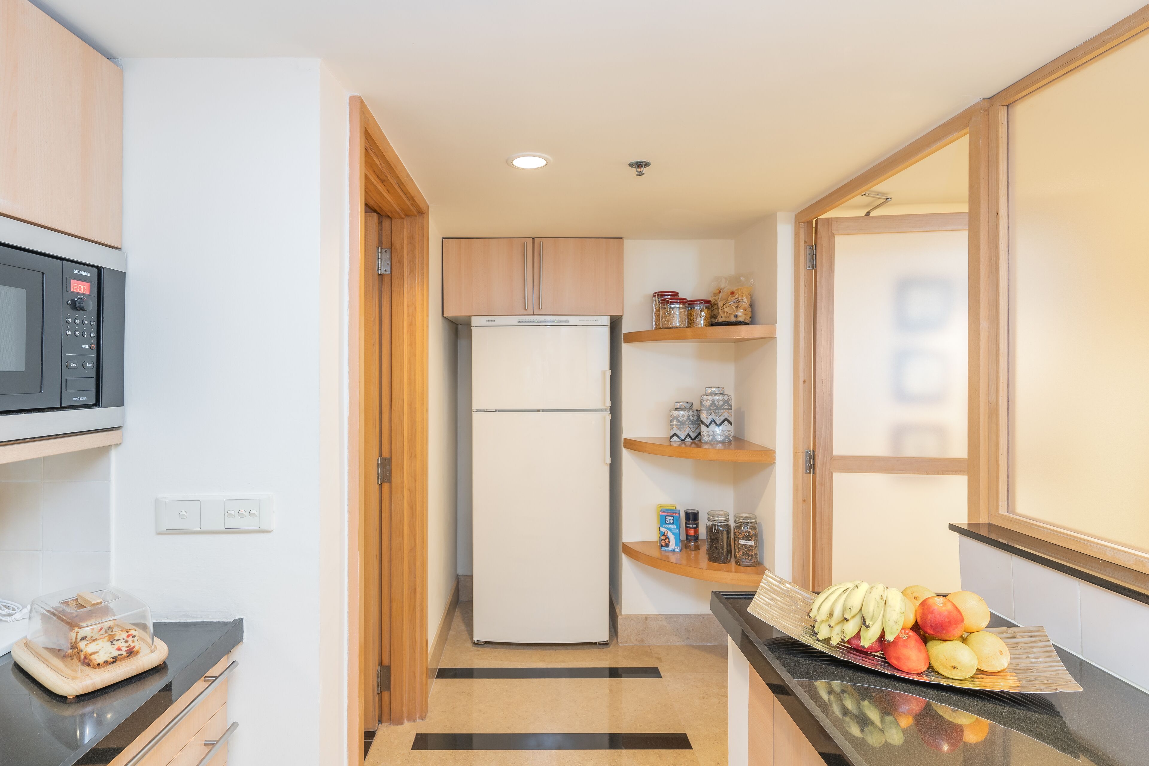A compact kitchen with a microwave, refrigerator, wooden cabinets, and a fruit display.