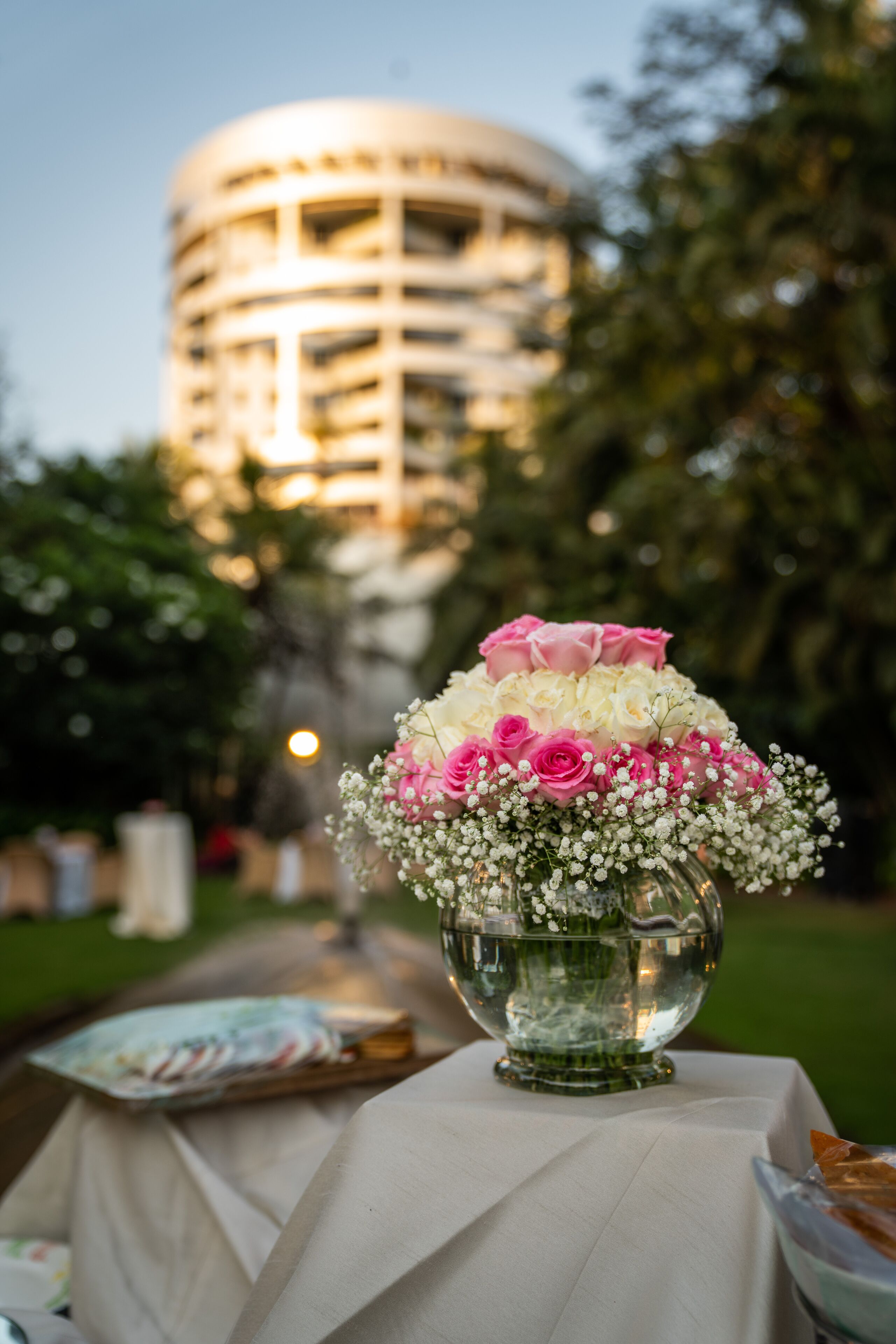 A table with a floral arrangement and a plate of food, set outdoors with a building in the background.