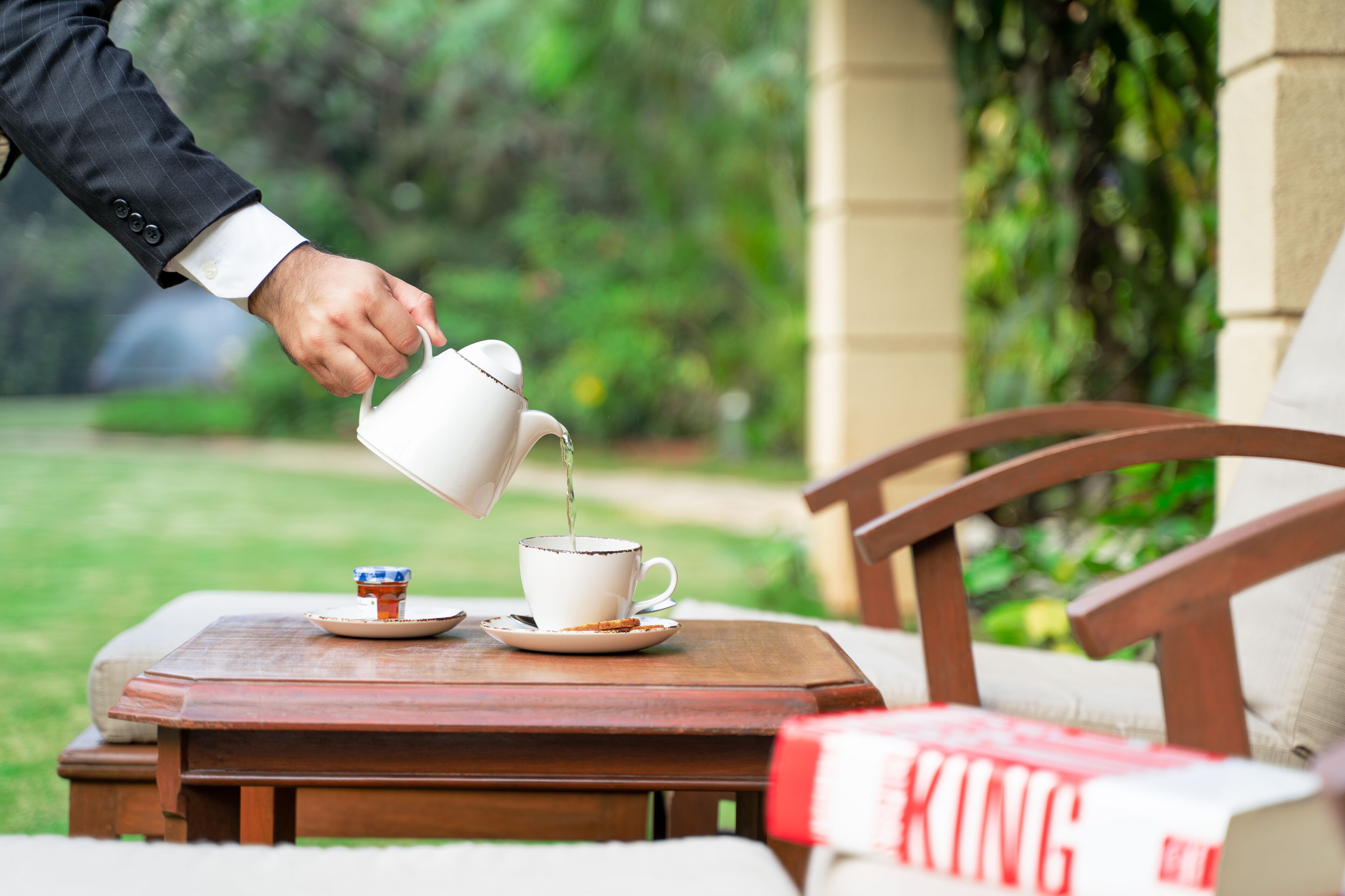 A hand pouring tea into a cup on a wooden table with a book titled "KING" beside it.